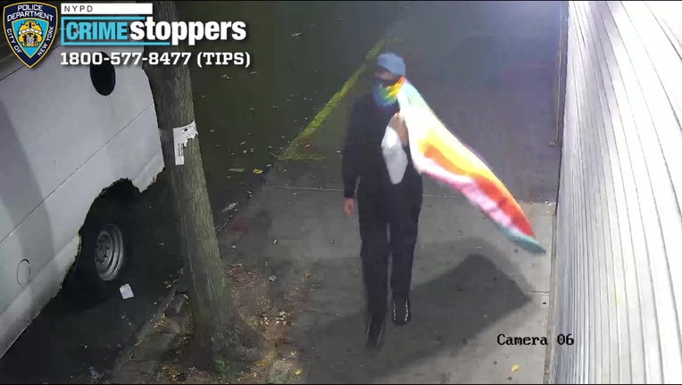 A person in all black, a dark had and dark sunglasses walking down a sidewalk with a rainbow covering over their face and a Pride flag draped over their shoulder.