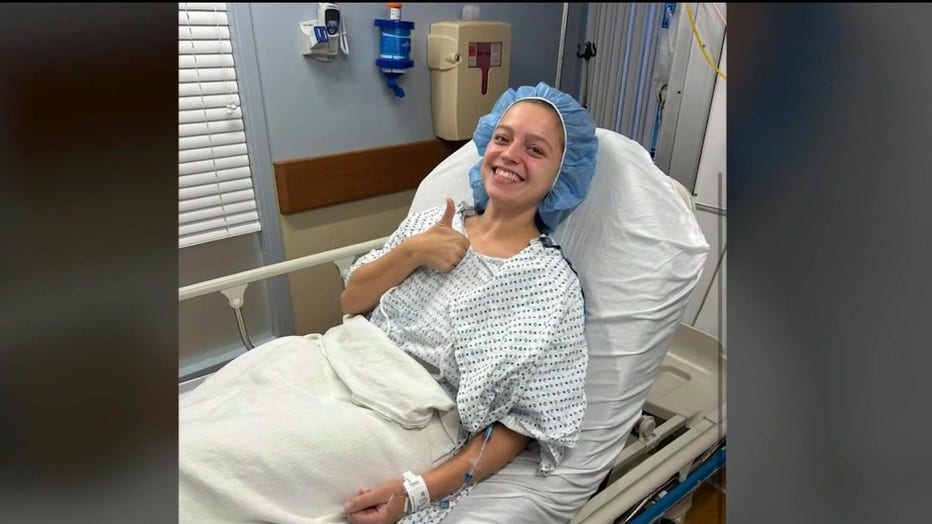 A woman on a hospital gurney sitting up, smiling and giving a thumbs up to the camera.