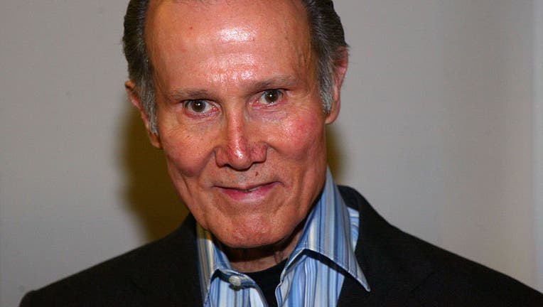 Photo of actor Henry Silva, wearing a blue striped shirt and black blazer in front of a gray background
