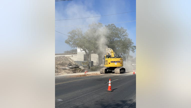 Photo of a gas plume coming from a gas line that was struck by a yellow Komatsu excavator in Southeast Austin. the plume is light gray against two green trees in front of a gray building and next to dirt and construction debris.