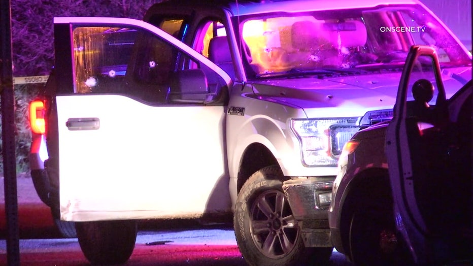 A white pickuip truck with the passenger door open, seen with bullet holes. Police lights can be seen reflected off the truck.