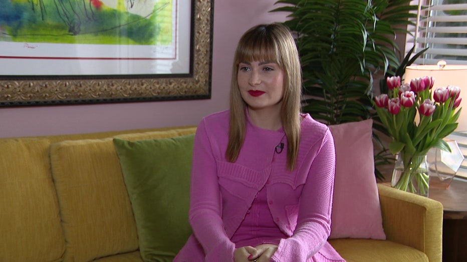 Molly Burke is sitting on a couch in a colorful living room wearing a pink dress. She is wearing red lipstick and has a soft smile on her face. Molly has shoulder-length straight hair with bangs.