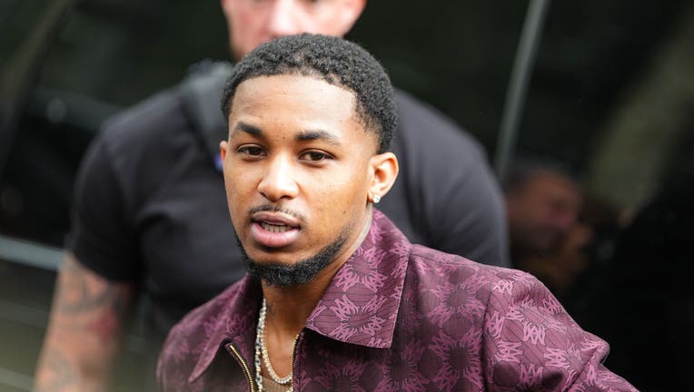 DDG wears a burgundy jacket featuring a subtle, intricate pattern. The jacket has a pointed collar and a zip fastening. Underneath, a brown ribbed shirt is visible. Jewelry includes multiple layered necklaces with a prominent pendant and small stud earrings. Hair is styled in a short, textured cut, outside Amiri, during Paris Fashion Week - Menswear Spring/Summer 2026, on June 26, 2025 in Paris, France (Photo by Edward Berthelot/Getty Images)