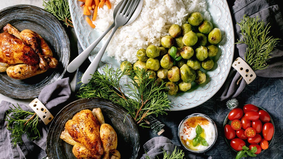 Christmas dinner table with grilled mini chicken, rice and vegetables baked brussel sprouts, baby carrot in ceramic bowls, xmas decorations on blue linen tablecloth over grey background. Flat lay