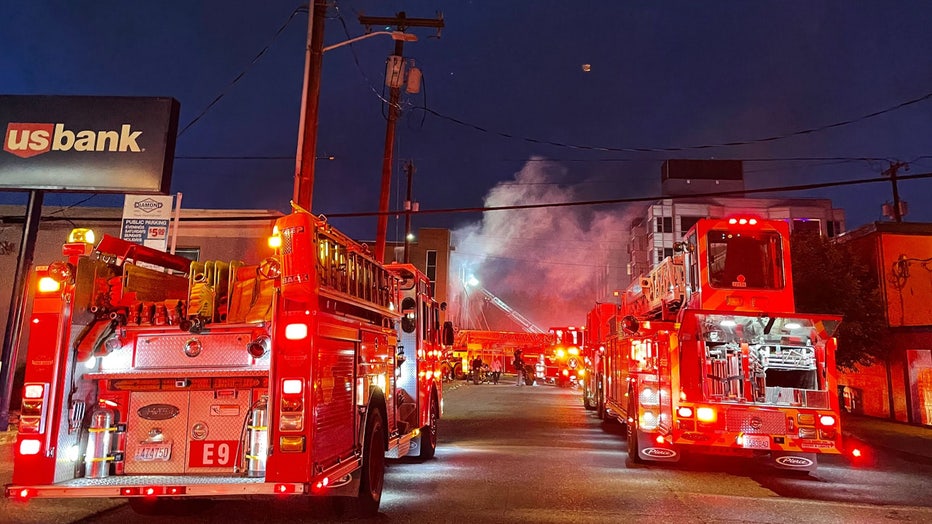 Multiple firetrucks with lights on parked outside of a burning building in Seattle. Smoke is scene coming out of the building and a rescue latter is visible.